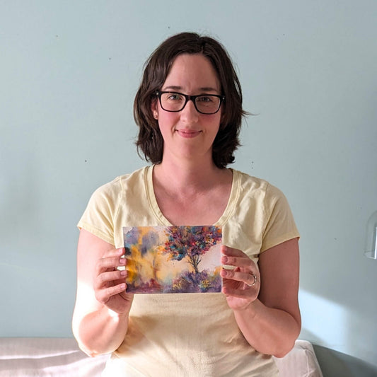 Woman holding a colorful painting with a tree design in front of a light blue wall.