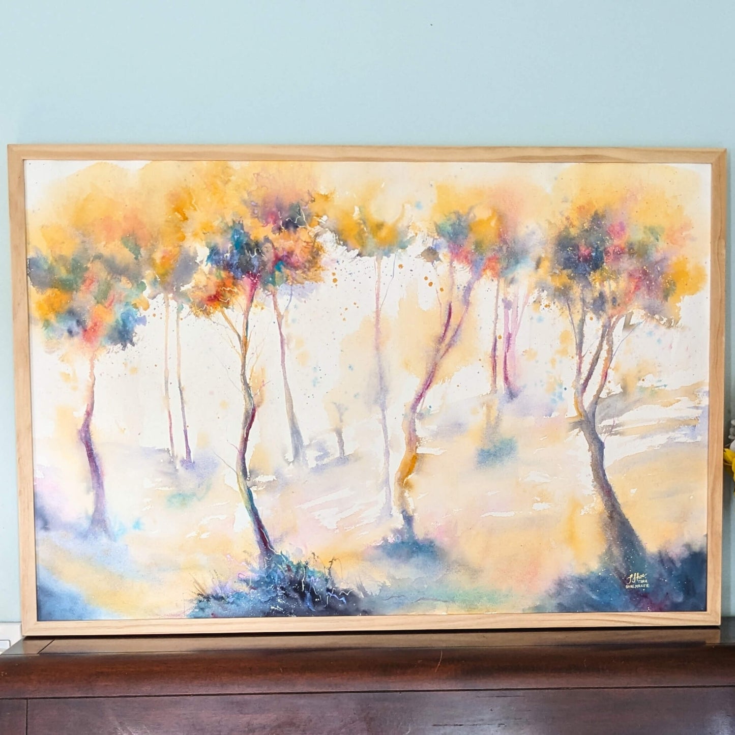 Framed abstract painting of trees with colorful brushstrokes on a light blue wall.