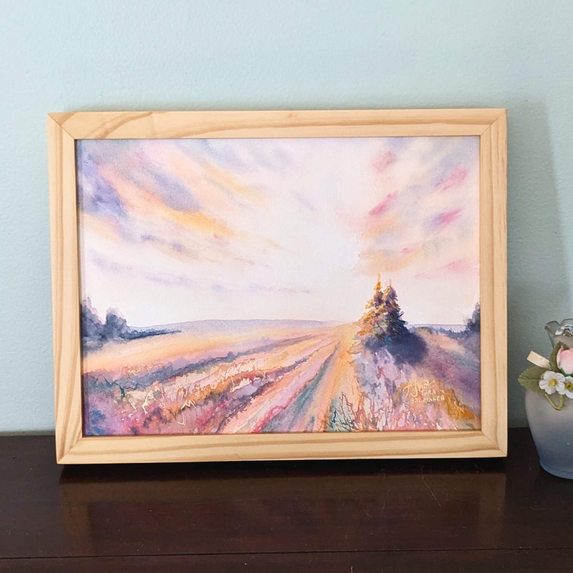 Framed painting of dawn over a field with a tree on a wooden surface.