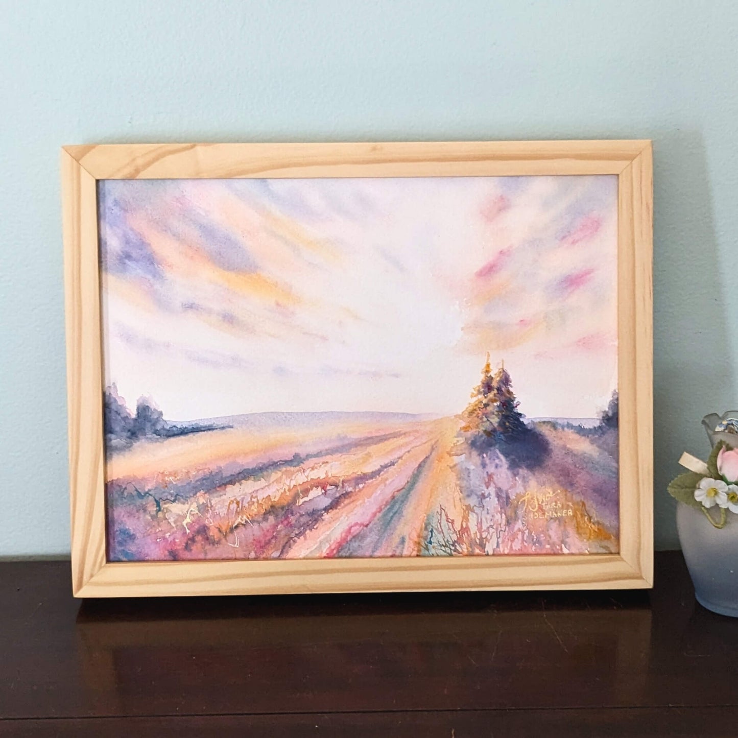 Framed painting of dawn over a field with a tree on a wooden surface.