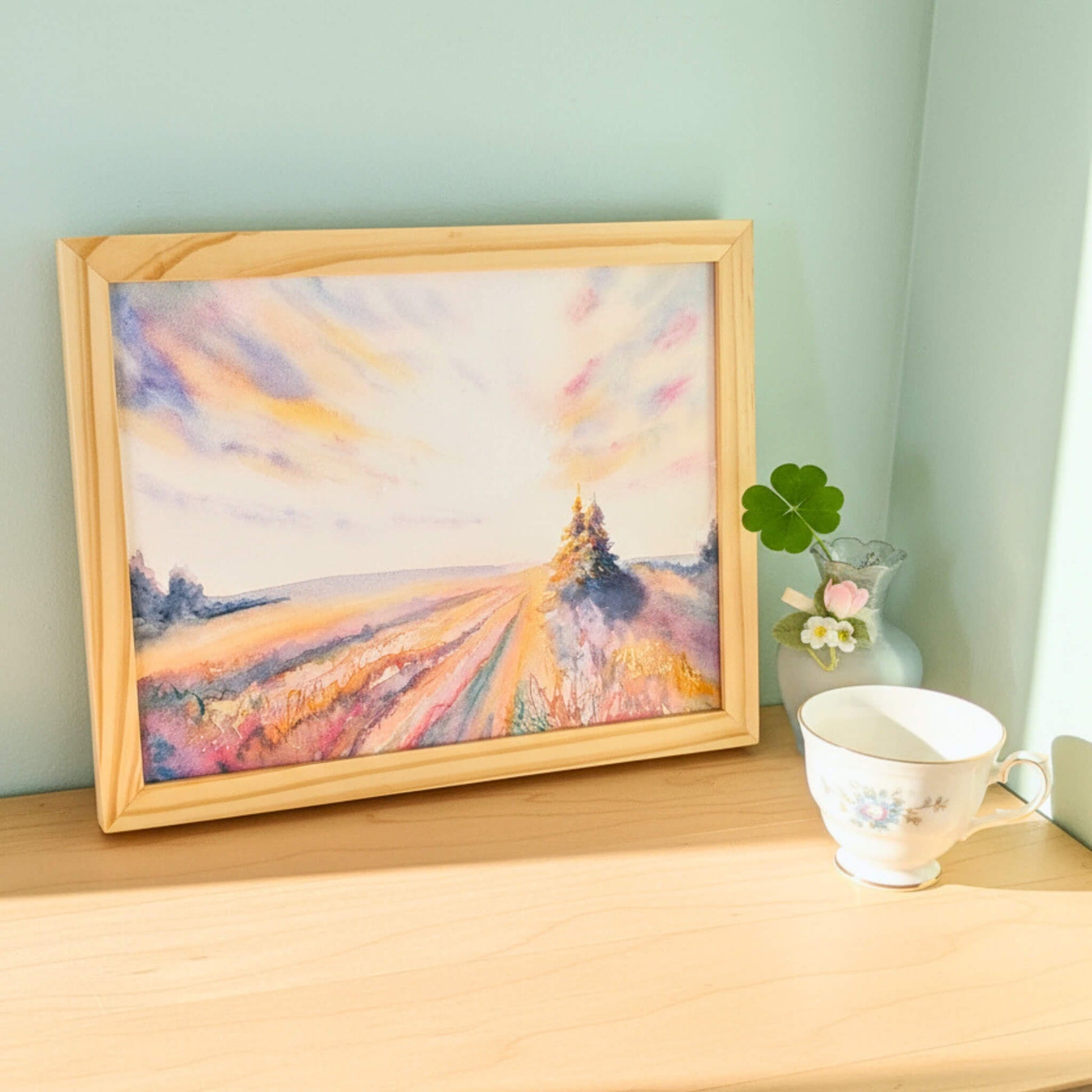 Framed painting of a landscape on a wooden surface with a cup and vase.