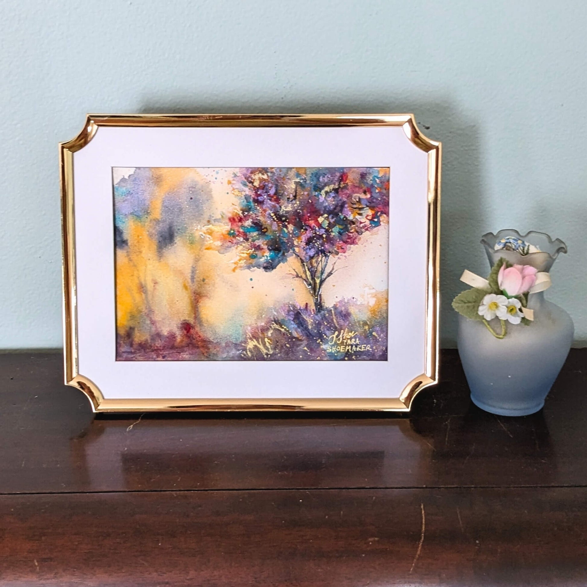 Framed abstract painting on a wooden surface with a small vase and flowers.