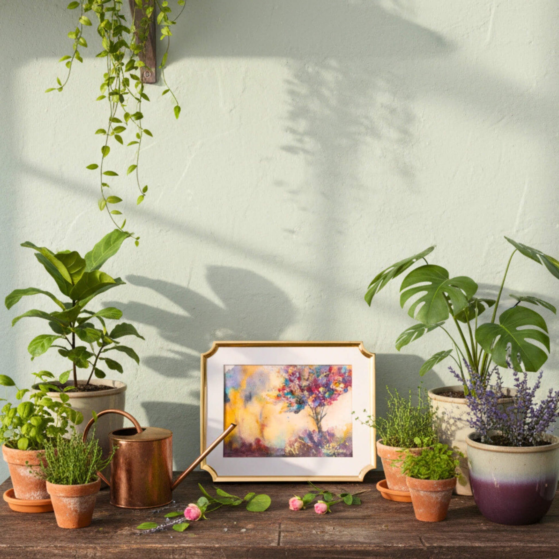 Decorative table with potted plants, a watering can, and a framed painting against a light wall.
