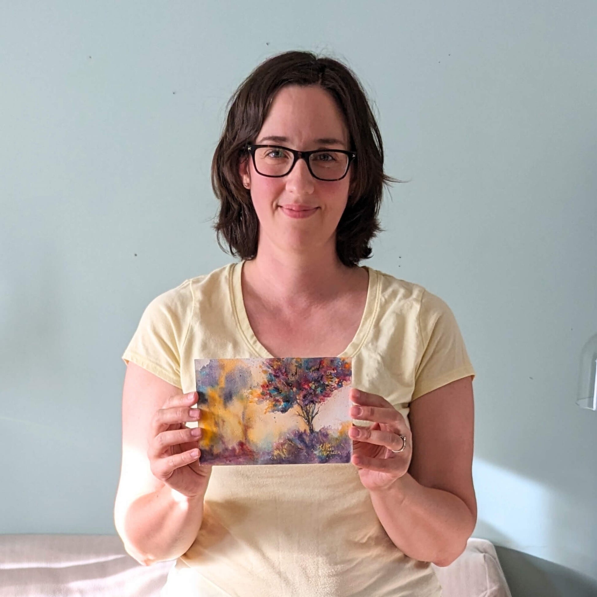 Woman holding a colorful painting with a tree design in front of a light blue wall.