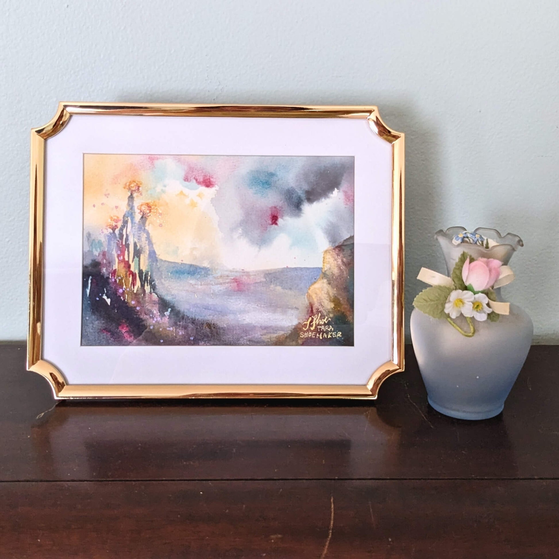 Framed artwork of three burning trees on a hill with a vase of flowers on a wooden surface.