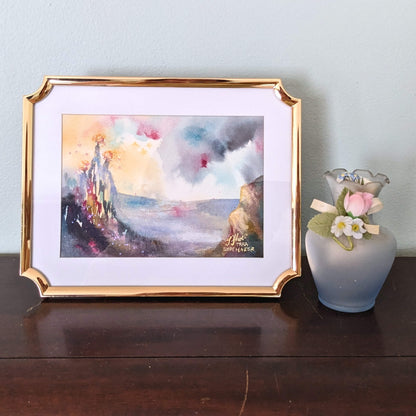 Framed artwork of three burning trees on a hill with a vase of flowers on a wooden surface.