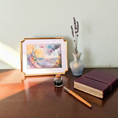 Framed artwork, inkwell, pen, and notebook on a wooden surface with a light background
