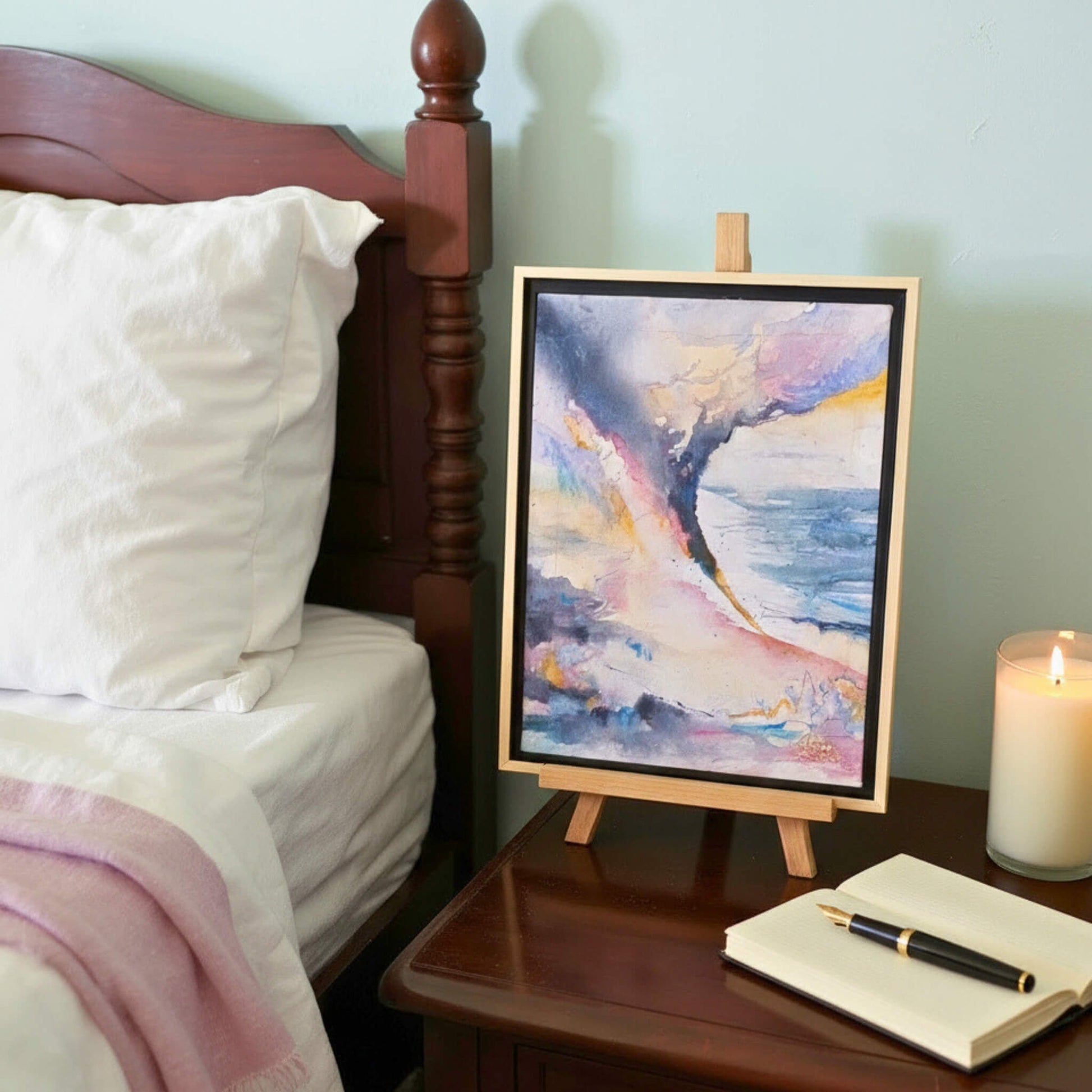 Colorful abstract painting on a small easel next to a bed with pillows and a candle.