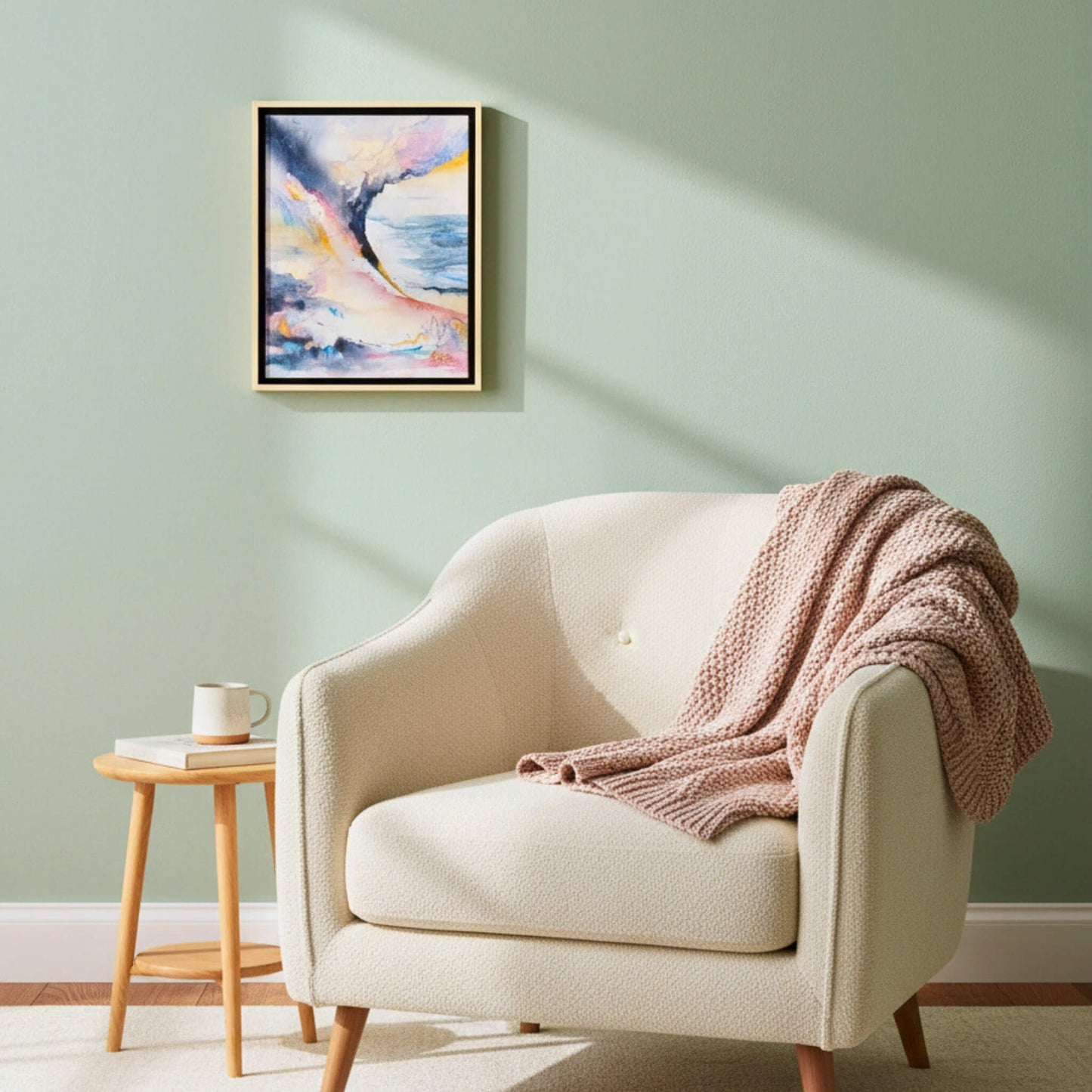 Beige armchair with a pink blanket in a room with a light green wall and a framed abstract painting.