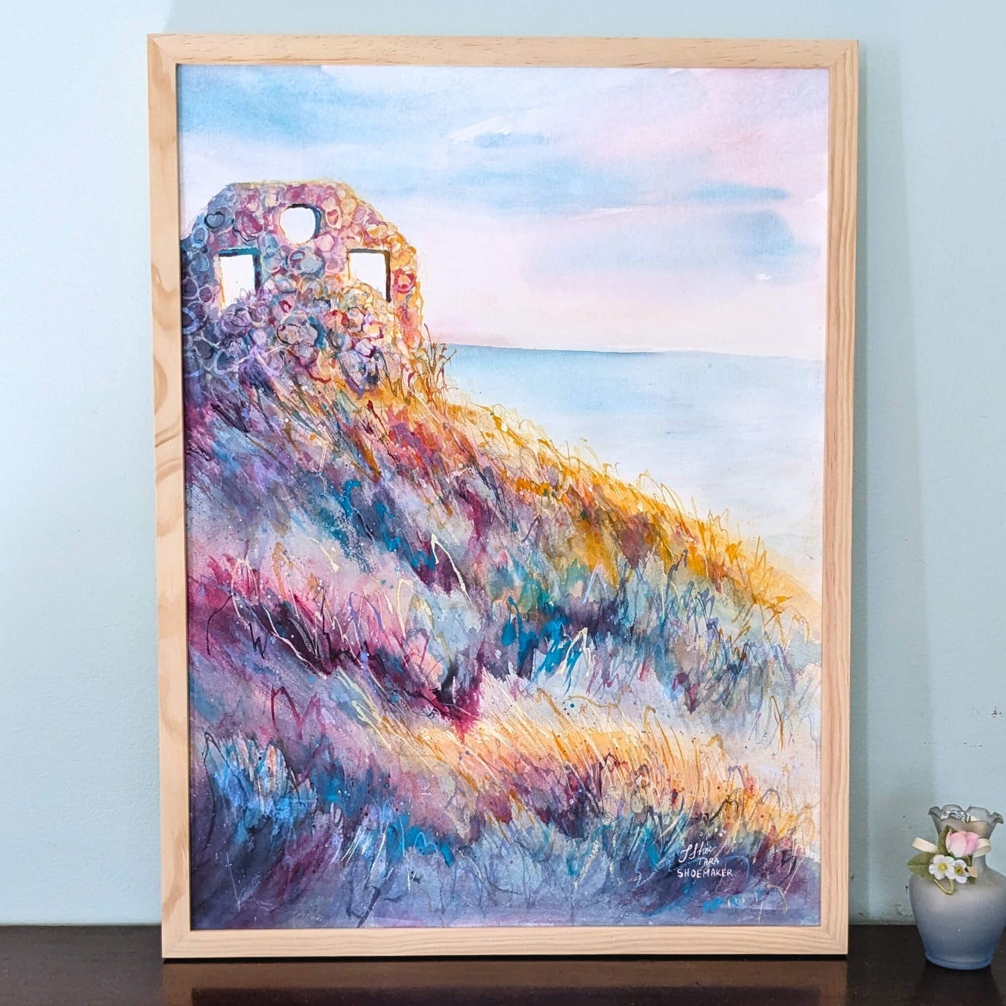 Framed colorful painting of a ruin on a hillside against a blue sky.