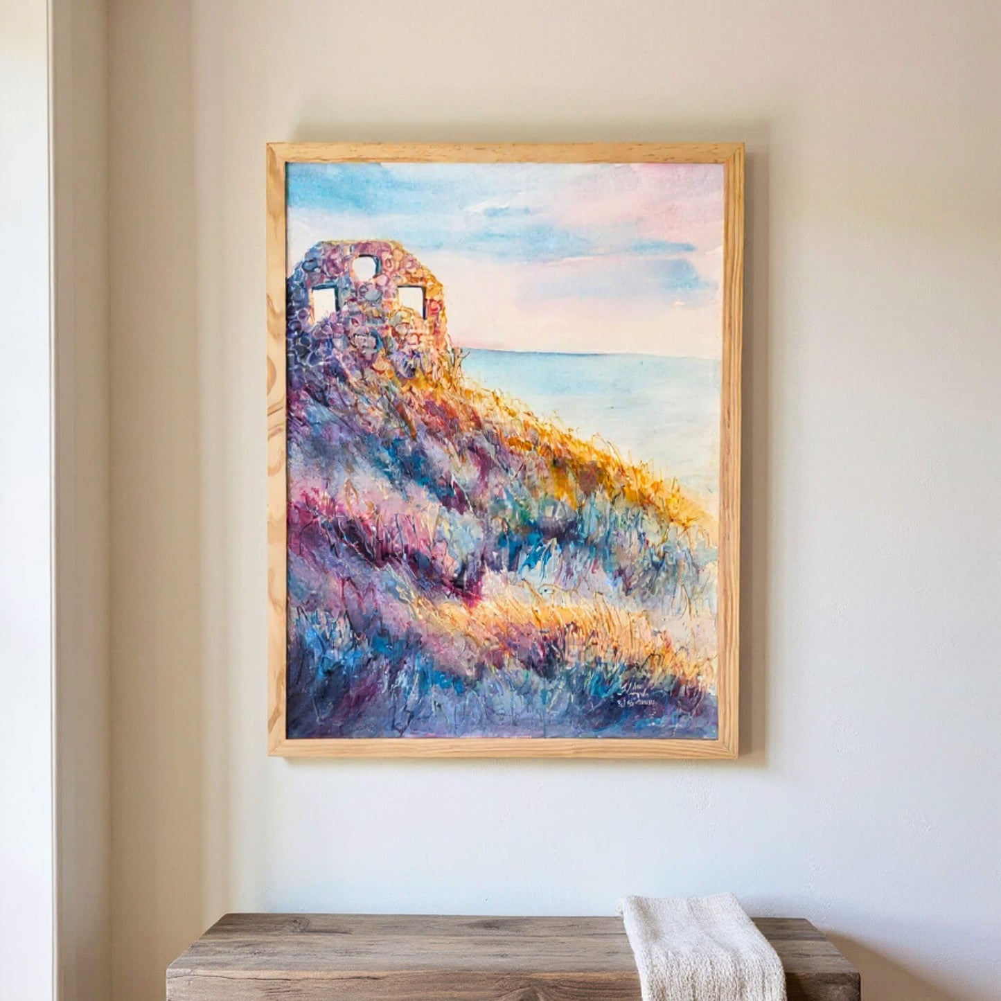 Framed painting of a colorful ruin above wild grasses on a wall above a wooden bench