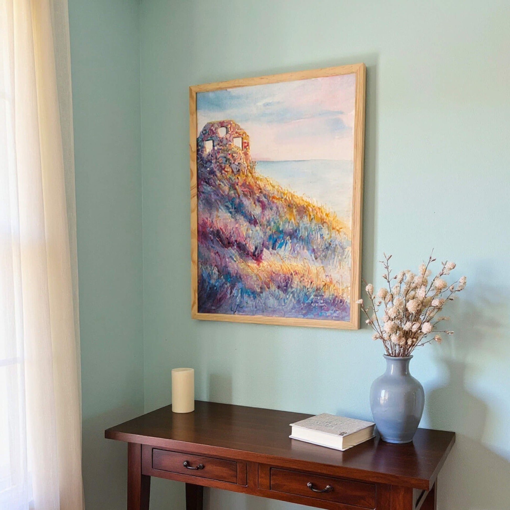 Framed abstract painting on a wall above a wooden desk with a vase and book.
