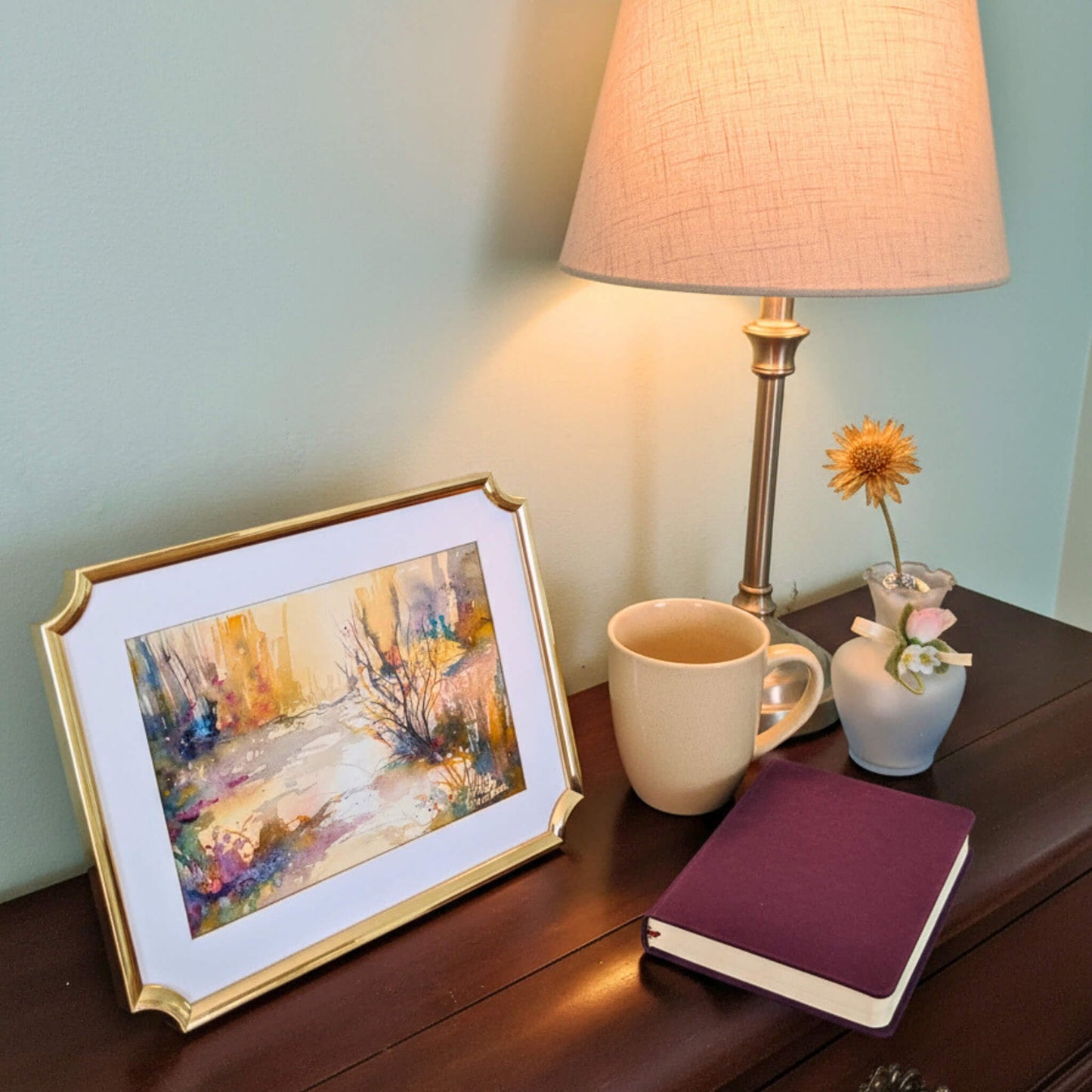 Framed painting, lamp, mug, and notebook on a wooden surface with a soft background.