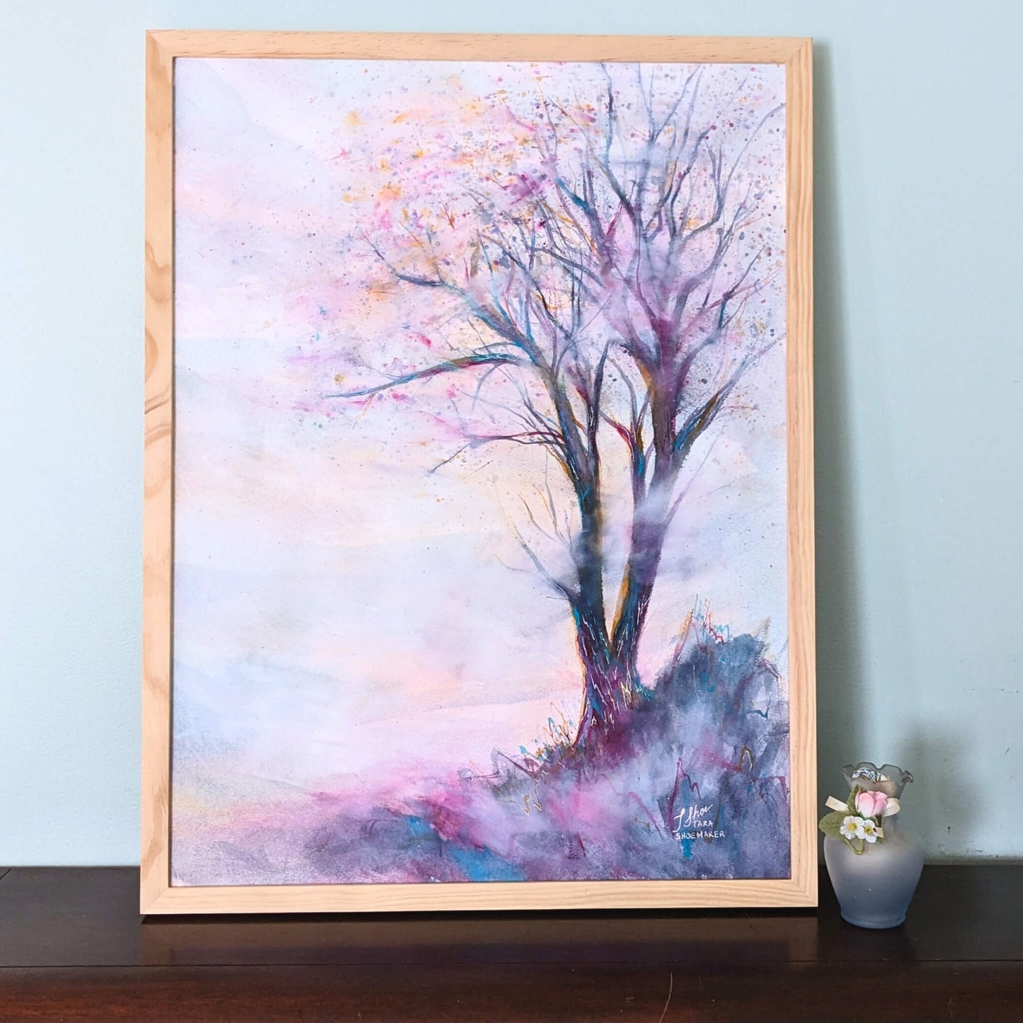 Framed artwork of a tree with pastel colors on a light blue wall.