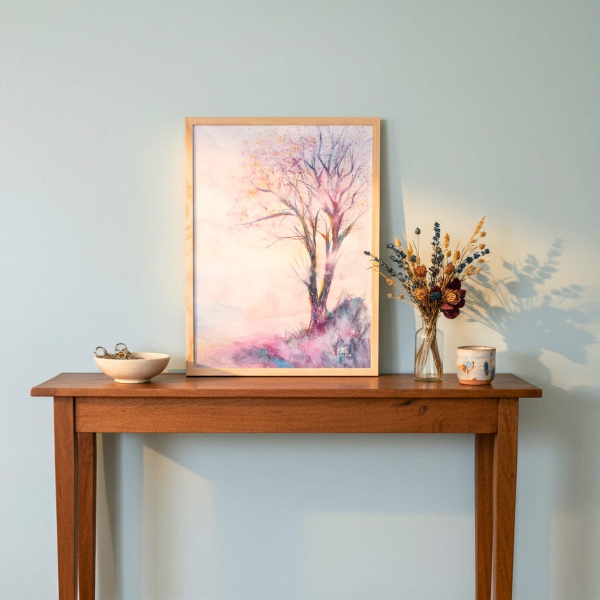 Wooden table with a framed artwork, vase with flowers, and small bowl against a light blue wall.