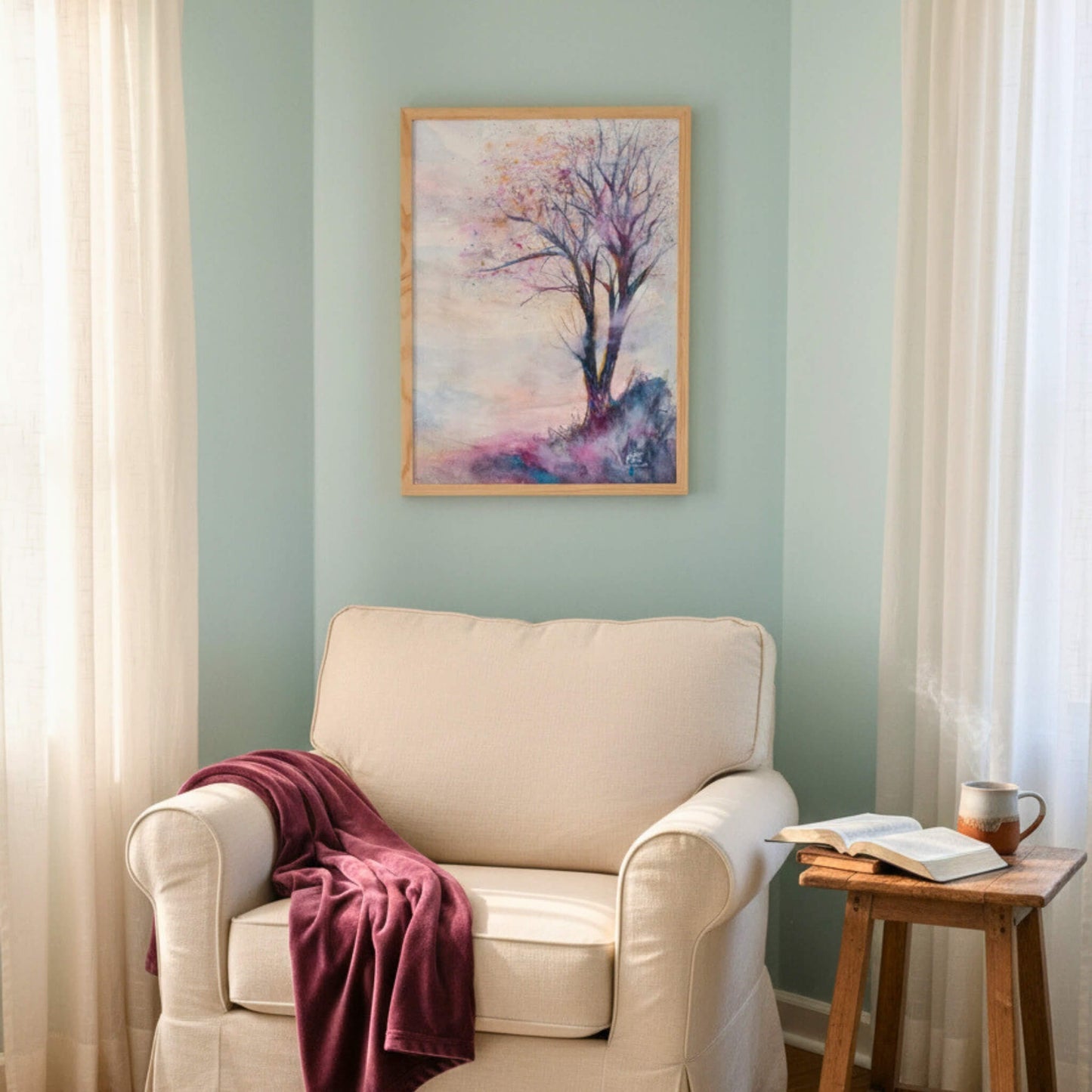Beige armchair with a red blanket, wooden side table with a book and mug, and a framed tree painting on a light green wall.