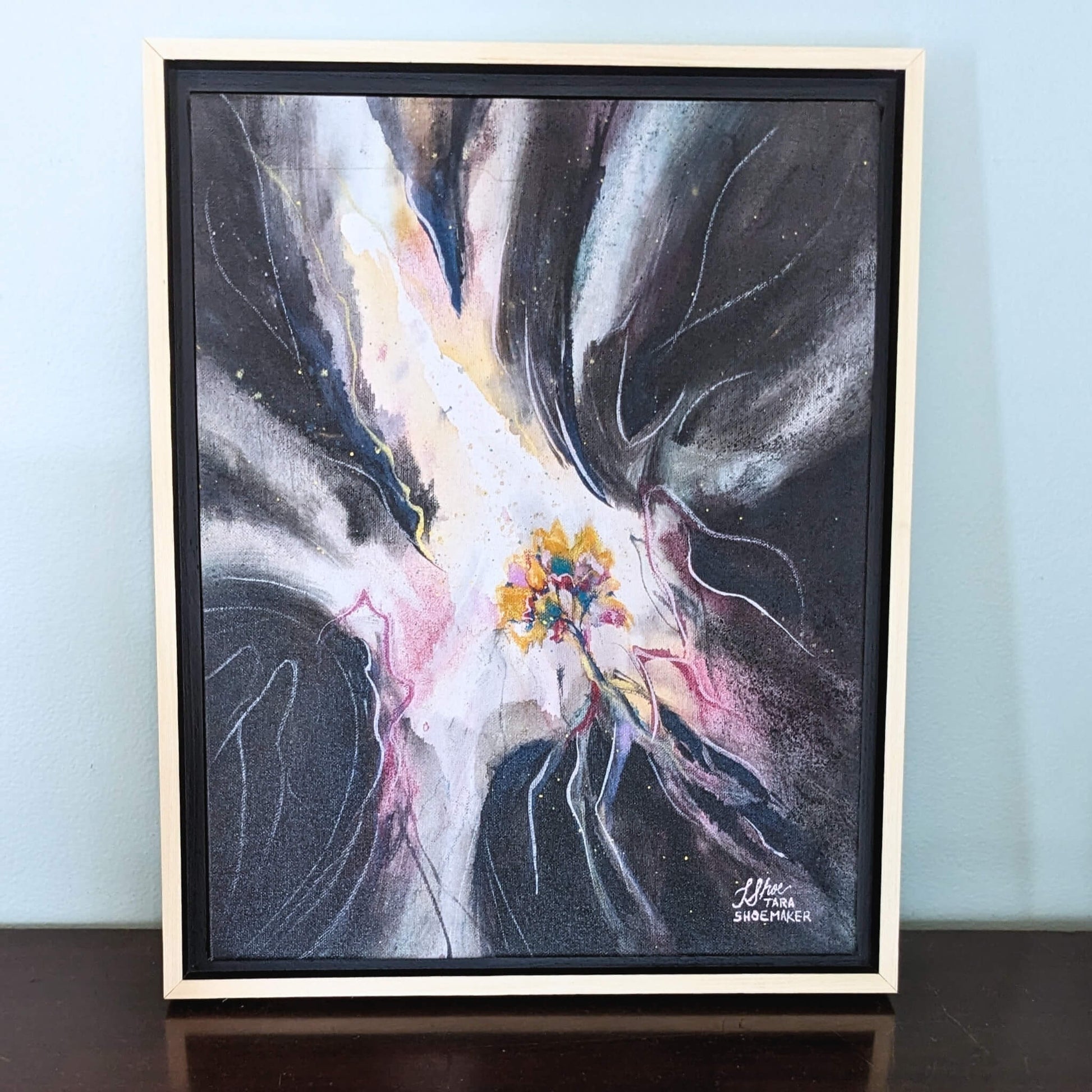 Framed abstract artwork with a central colorful tree and swirls of light and dark on a light blue wall