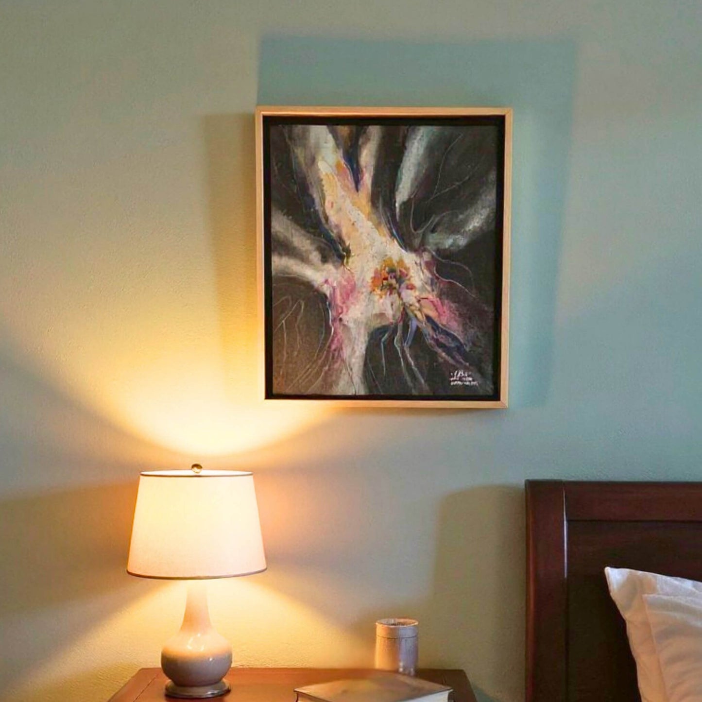 Framed abstract painting on a wall above a bedside table with a lamp and cup.
